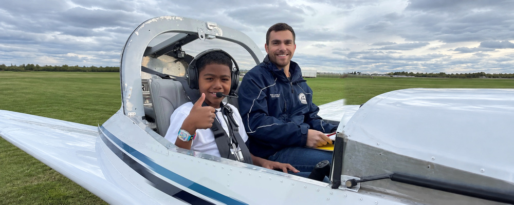 A smiling boy in a headset gives a thumbs up from the cockpit of a small plane beside his volunteer pilot