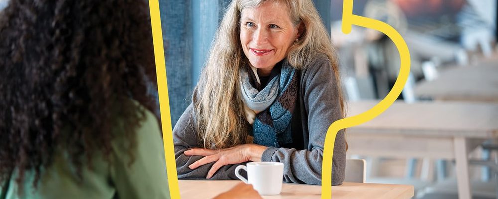 A woman smiling at a table with a white cup of coffee, framed by a yellow mug outline