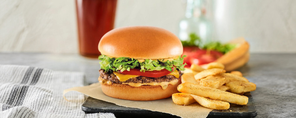 A cheeseburger with lettuce, tomato and pickles, with fries and a drink behind it